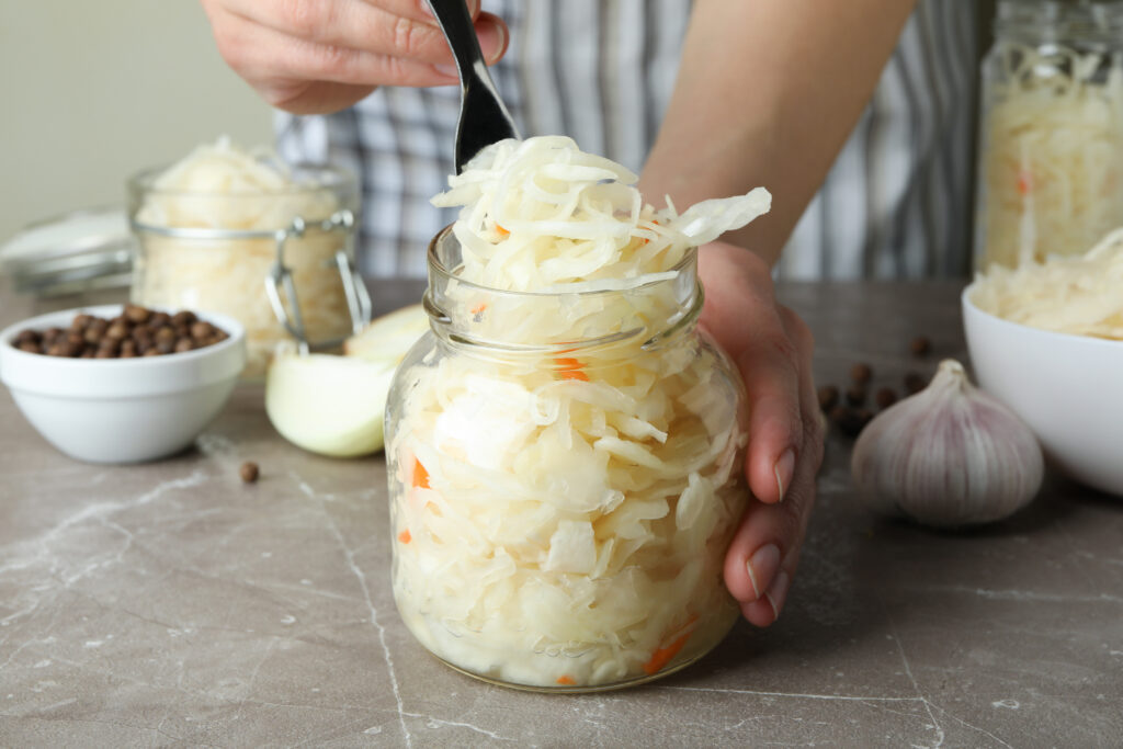 Woman holding jar with sauerkraut on gray textured table with ingredients